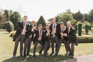 Groomsmen strike playful poses lined up outdoors in dark suits.