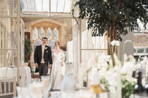 Elegant couple enters sunlit conservatory decorated with white flowers and greenery.