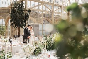 An elegant wedding reception setup in a glass conservatory with white floral arrangements and table settings.