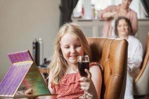 A child in a coral top holding a glass while sitting in a leather chair with others in background.