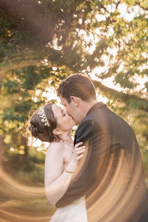 Romantic sunset wedding kiss with golden sunlight filtering through trees creating a dreamy atmosphere.