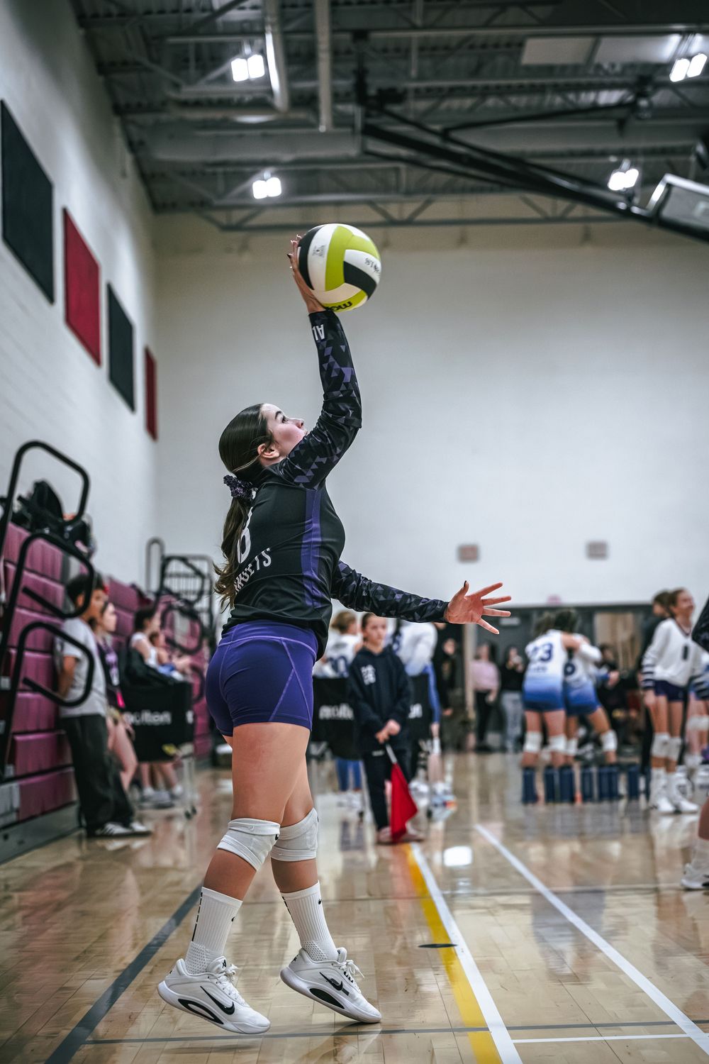 Female volleyball player in dark uniform leaps to serve the ball during an indoor gymnasium match.