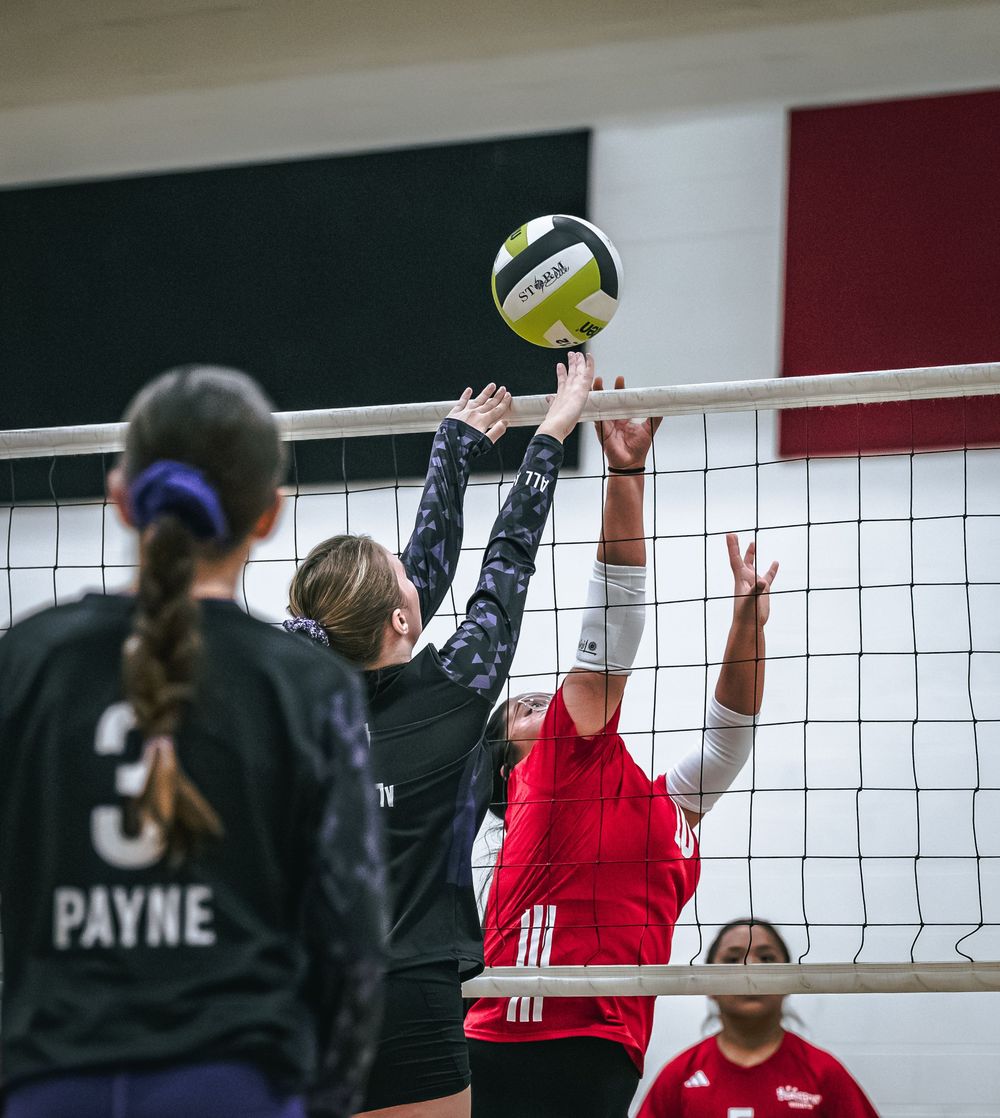 Two volleyball players battle at the net as player Payne watches during an indoor match.