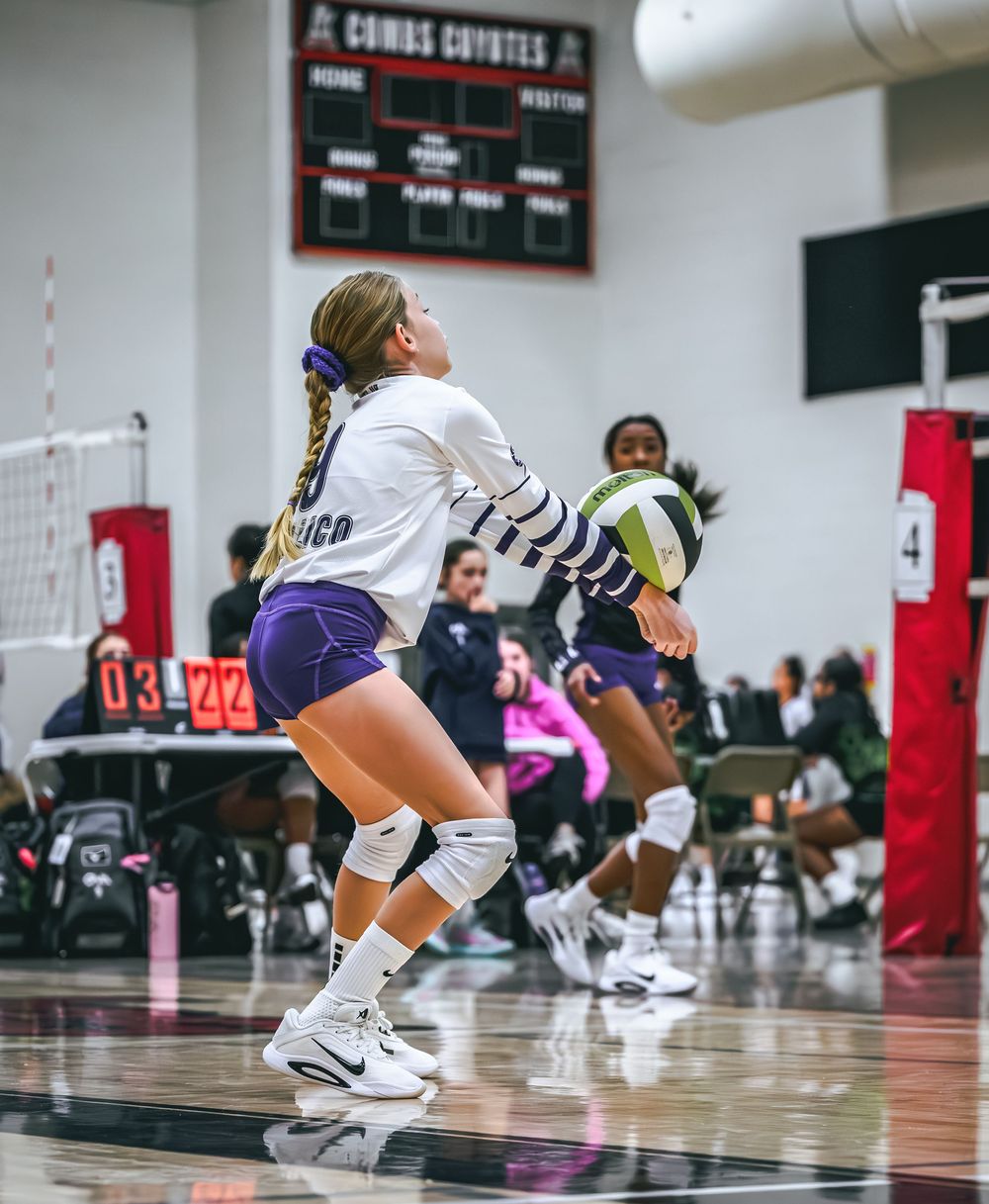 A blonde volleyball player in purple and white uniform bumps the ball during an indoor game.