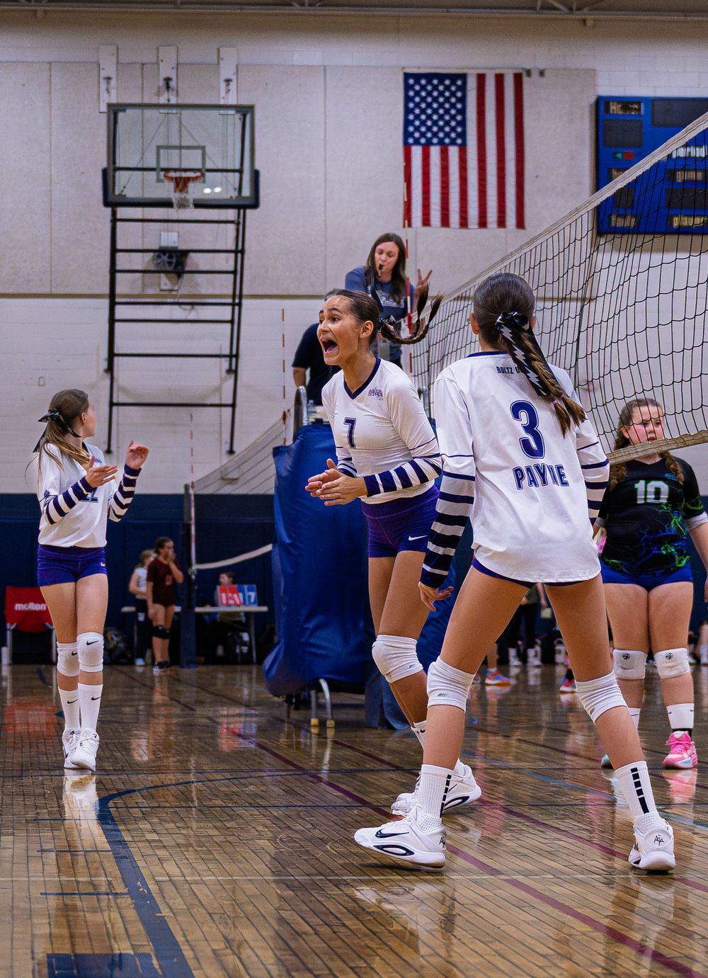 Female volleyball players in white uniforms celebrating a point during an indoor volleyball match.