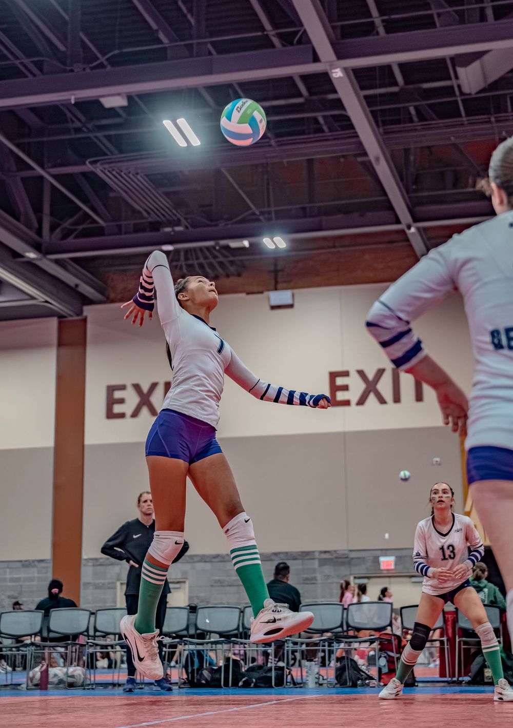 Female volleyball player leaping to spike the ball during an indoor tournament match with teammates visible.