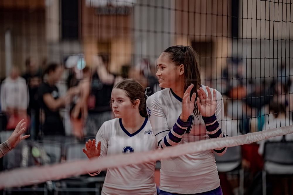 Two young female volleyball players in white uniforms stand at the net during an indoor volleyball match.