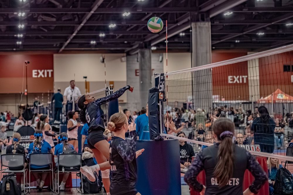 Indoor volleyball tournament with players spiking at the net in a large convention center gymnasium.