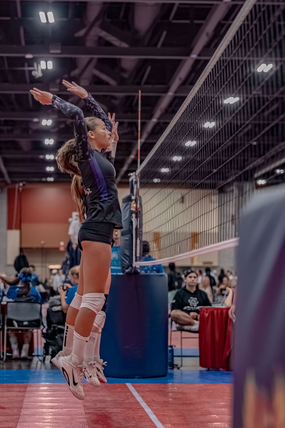 Female volleyball player leaps high near the net to spike or block during a competitive indoor volleyball match.