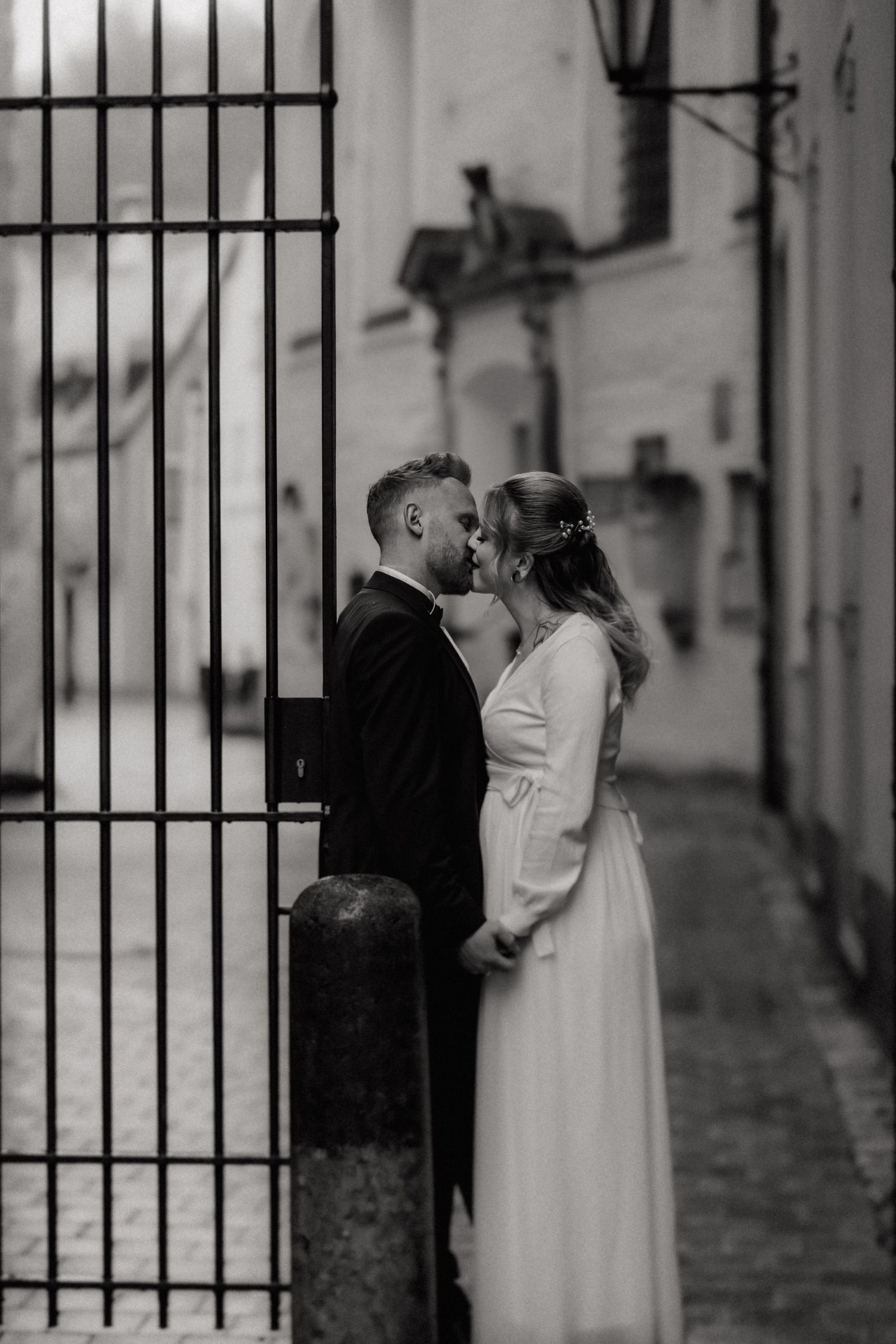 Black and white photo of a bride and groom sharing a kiss beside an iron gate on a cobblestone street.