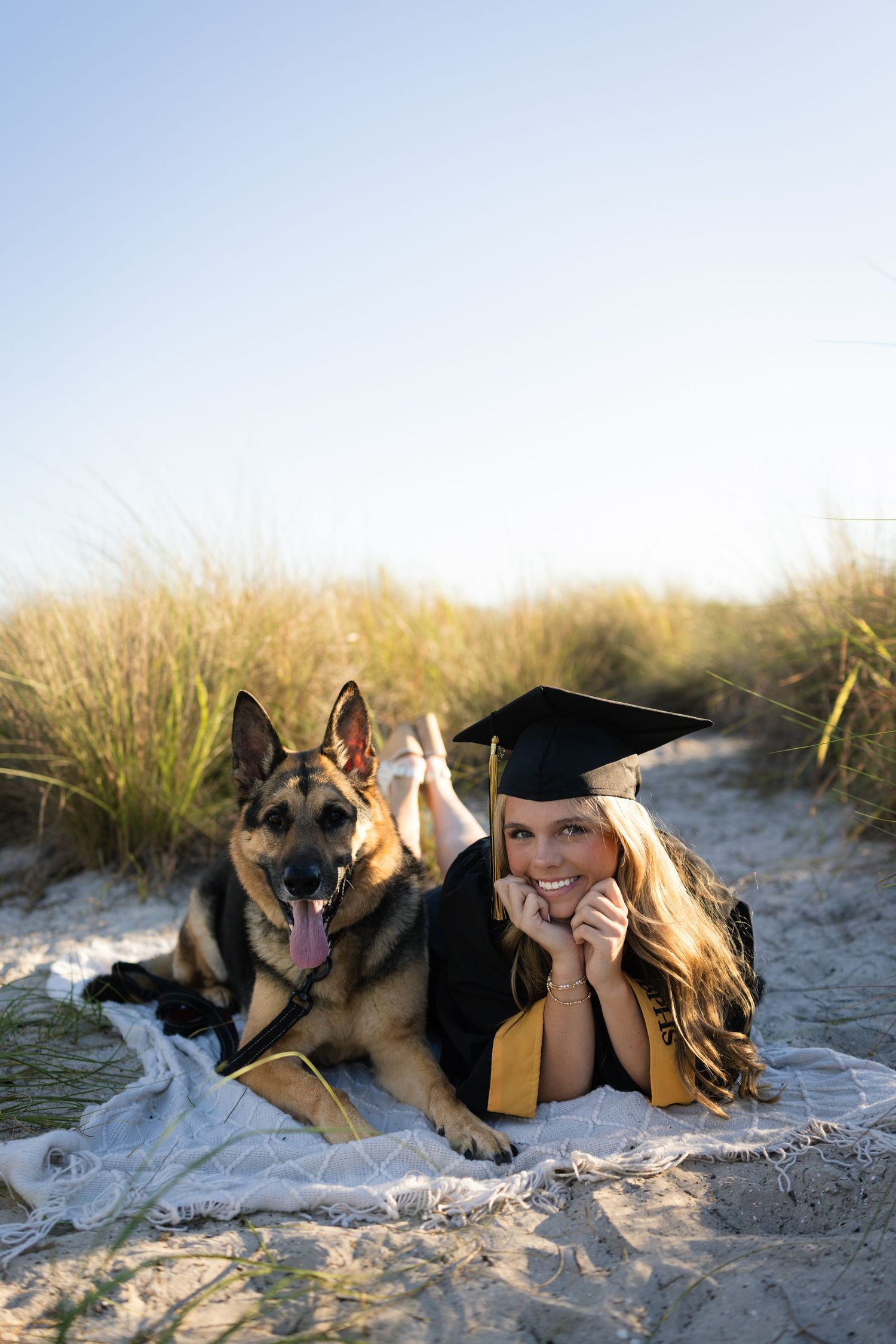 A smiling graduate in cap and gown lies on a beach blanket beside a German Shepherd dog among sandy dunes.