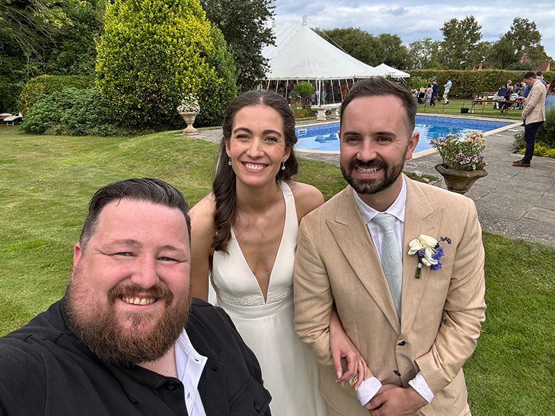 Bride and groom pose with a guest for a selfie at an outdoor wedding reception near a pool.