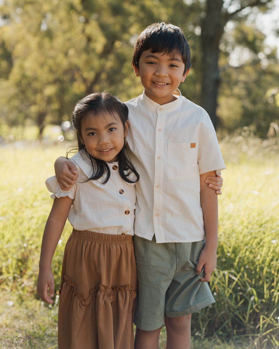 Two smiling children pose outdoors in sunny park, wearing earth-tone outfits, with the older boy hugging the younger girl family photoshoot