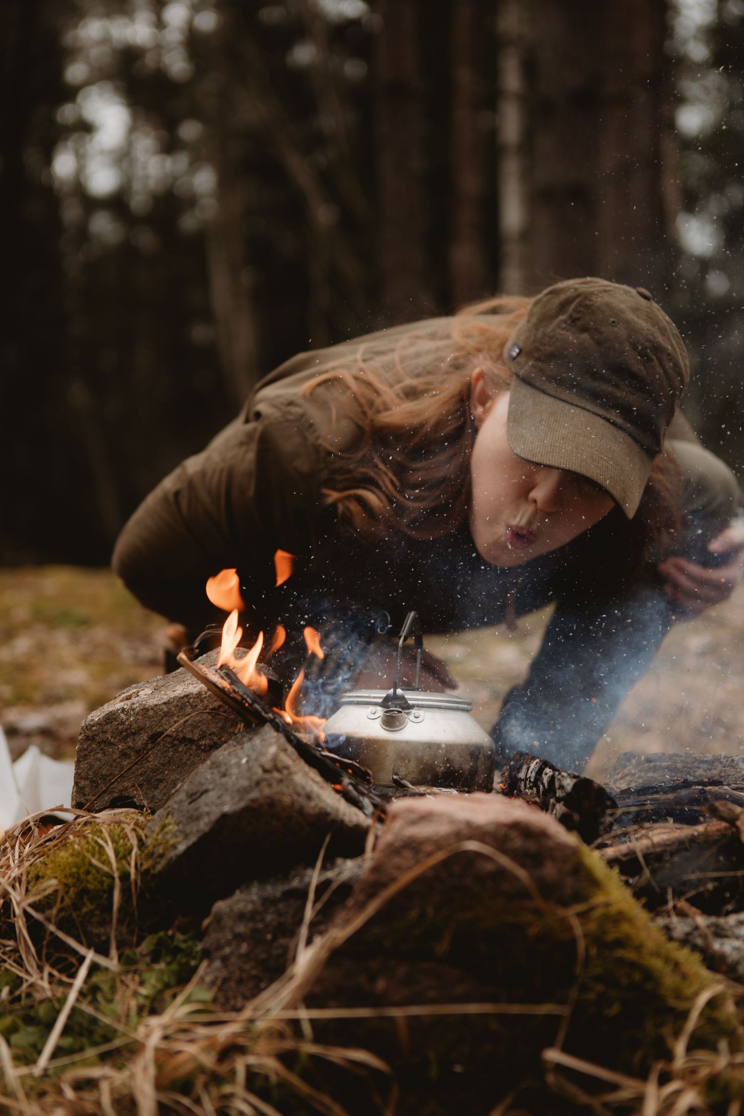 Woman in cap blowing on campfire with kettle on rocks in forest wilderness survival outdoor cooking scene.