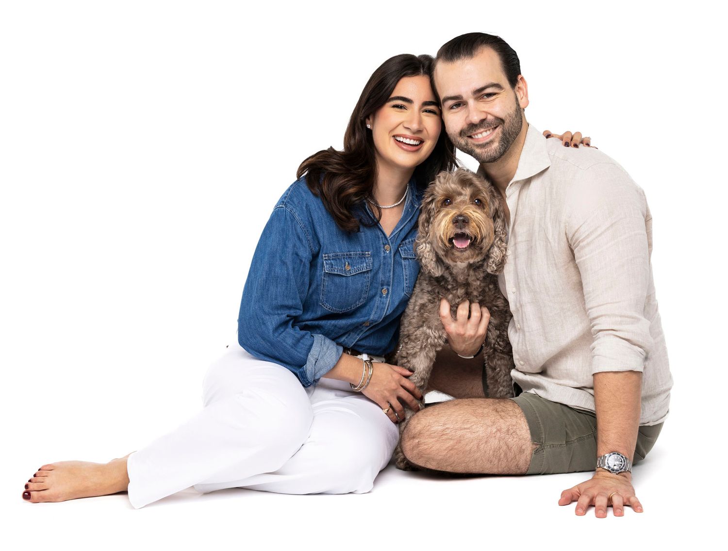 Smiling couple sitting on floor with fluffy brown dog, woman in blue denim shirt and white pants, man in beige shirt.