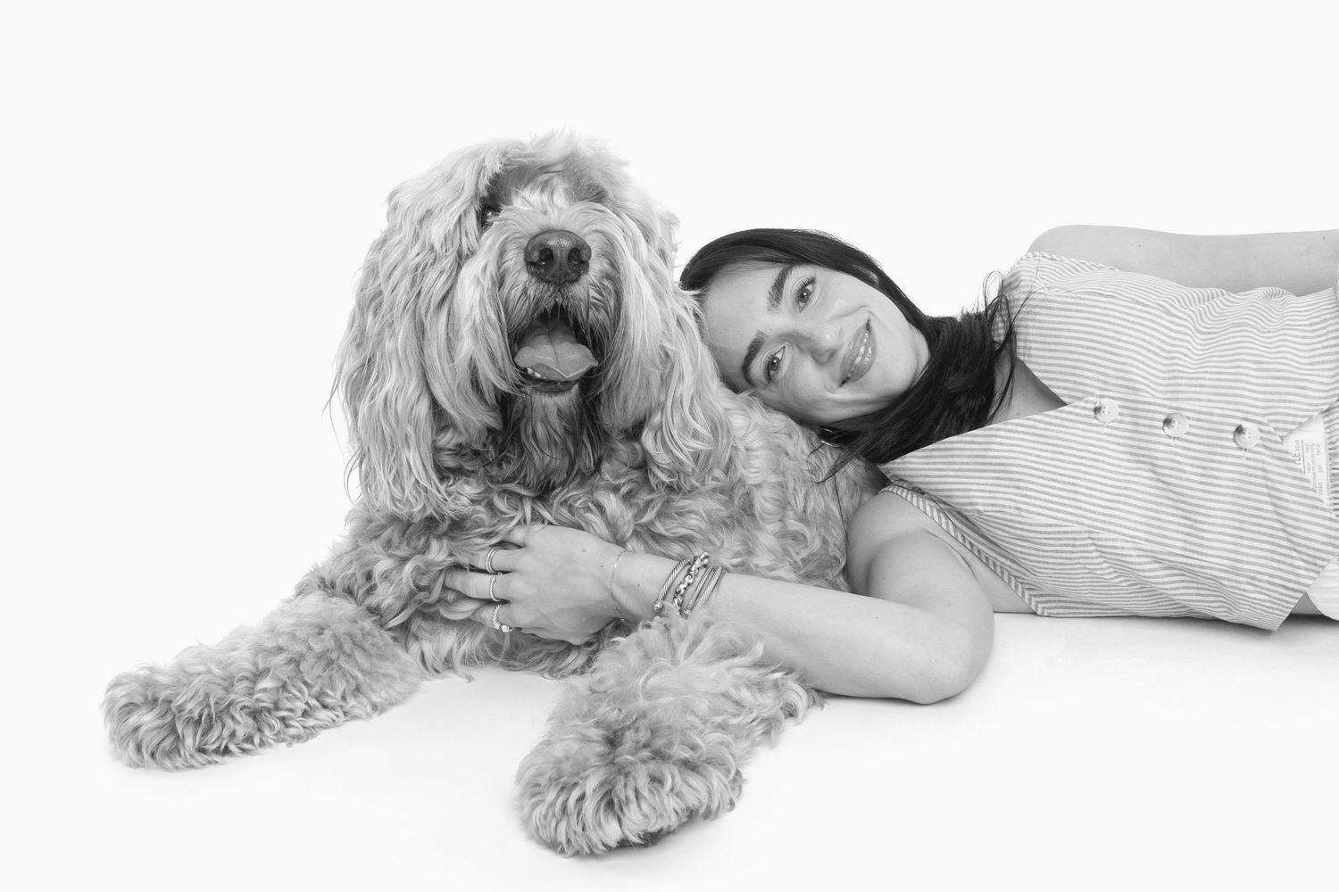 Black and white photo of a smiling woman lying beside a fluffy Goldendoodle dog on a white background.