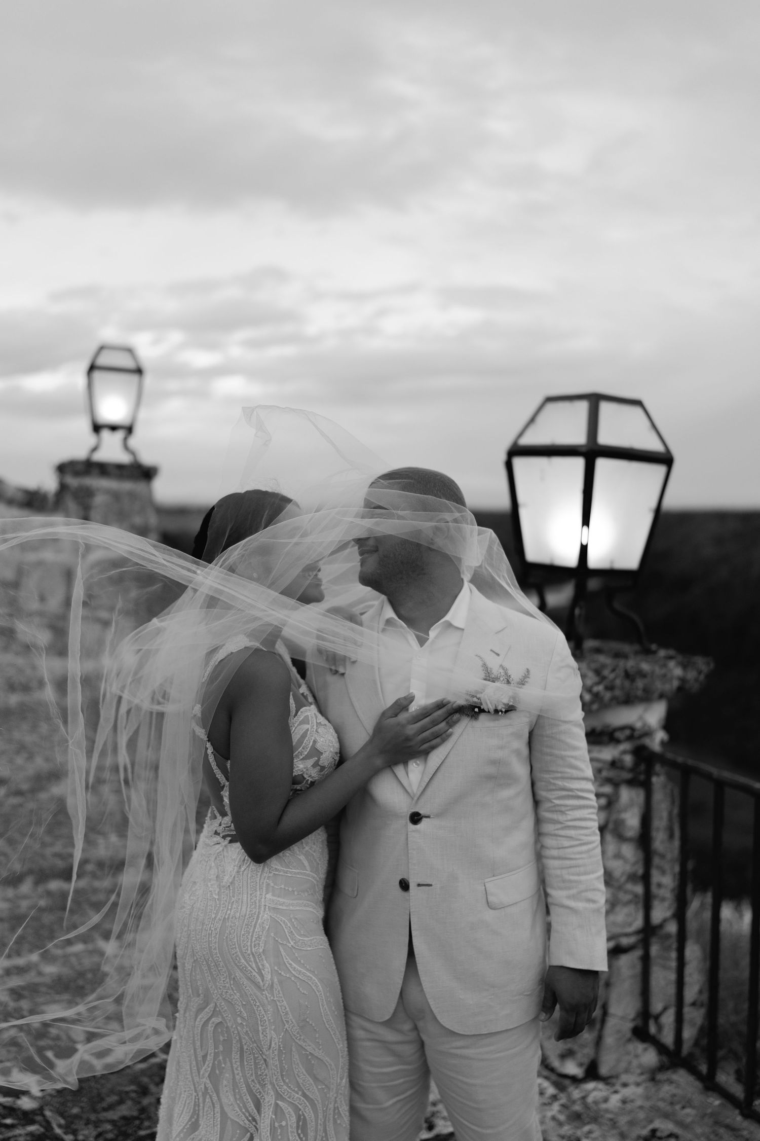 Couple in red dress and black suit embracing romantically before rustic stone building with wooden door.