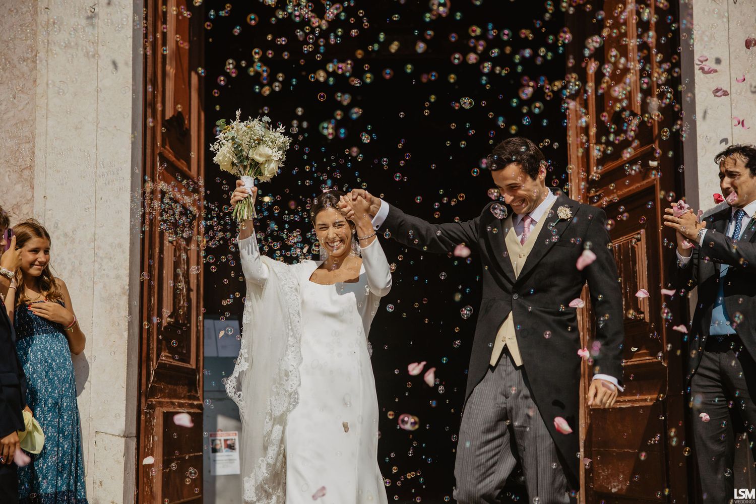 Joyful newlyweds exit church doors as colorful confetti fills the air, bride holding bouquet aloft in celebration.