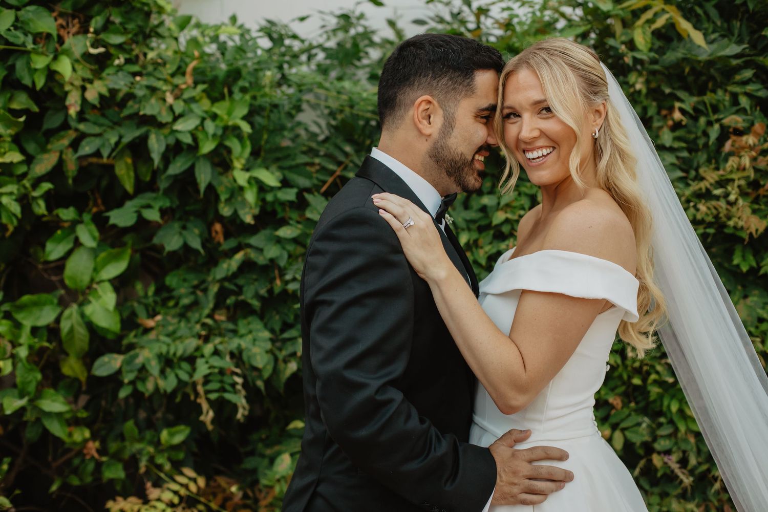 A smiling bride in an off-shoulder gown and groom in a black tuxedo share a romantic moment outdoors.