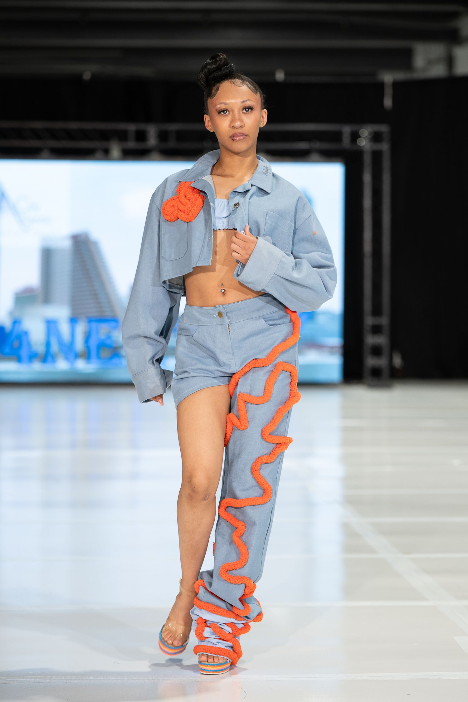 Model walks runway in denim crop set with orange ruffle trim pants at fashion show.