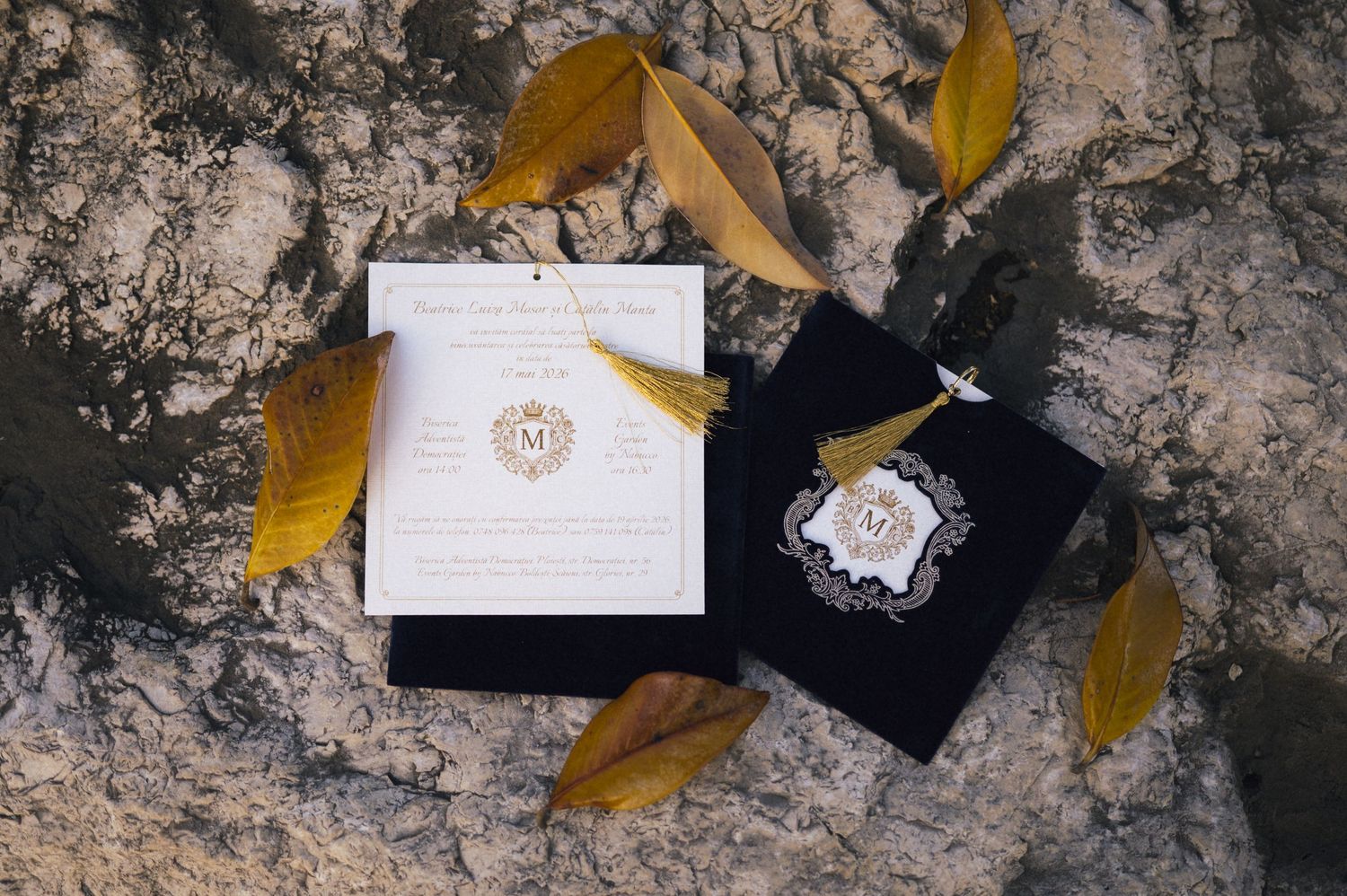 Elegant wedding invitation suite with gold monogram crest and tassel on rocky surface with autumn leaves.