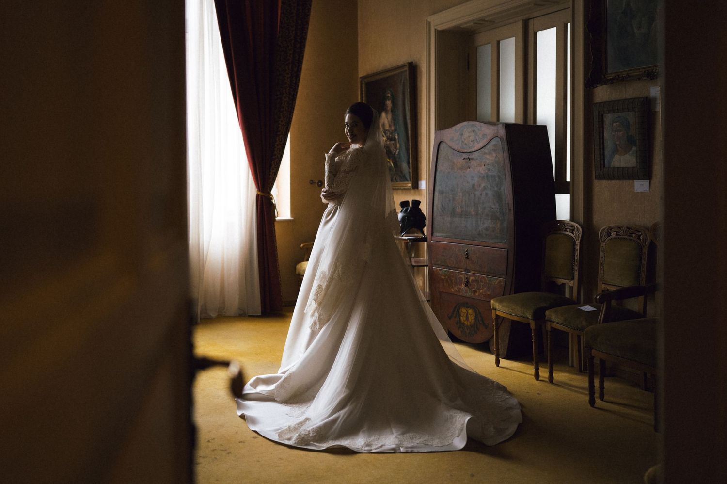 Bride in white wedding gown with long train stands by window in elegant room with vintage furniture.