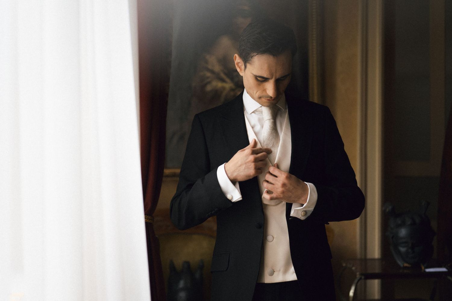 Dapper groom in black tuxedo adjusting his white cravat near a curtained window before wedding.