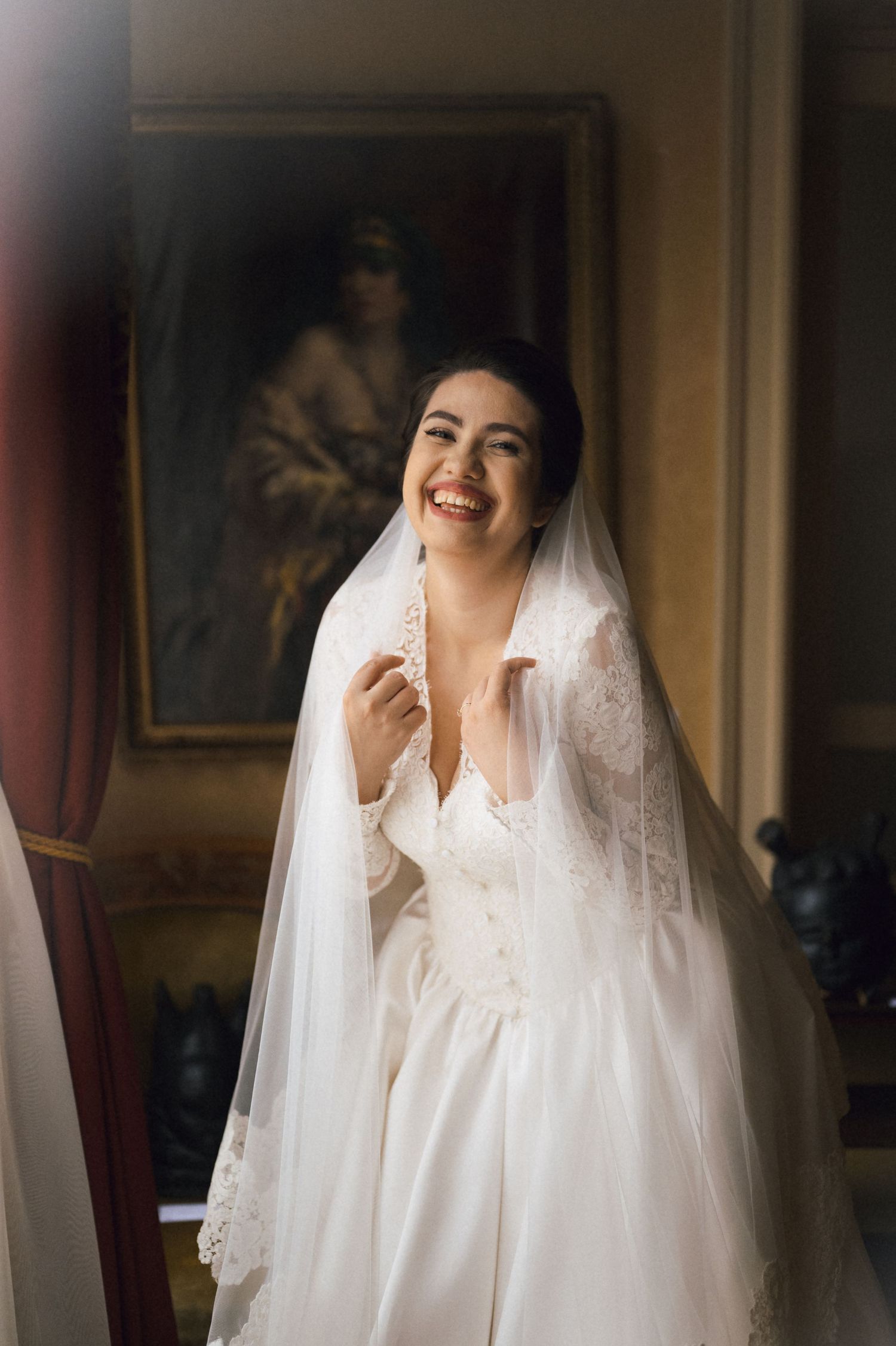 Smiling bride in white lace wedding dress and veil standing in elegant room with classic portrait painting.