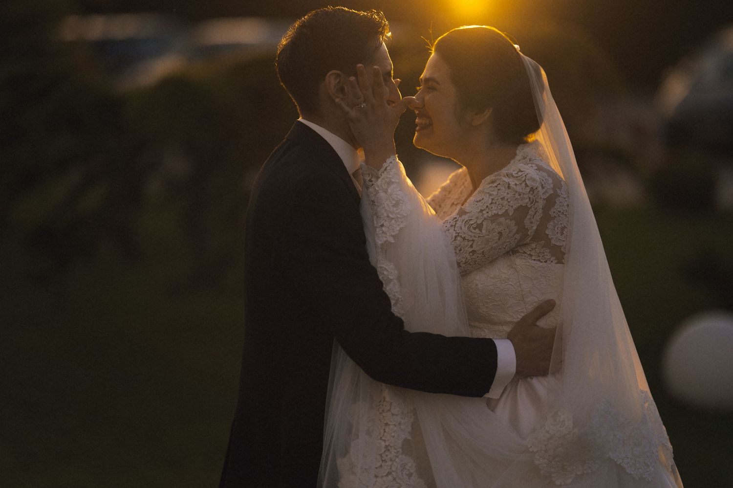 A bride and groom share a romantic kiss at sunset, with golden backlight illuminating their wedding attire outdoors.