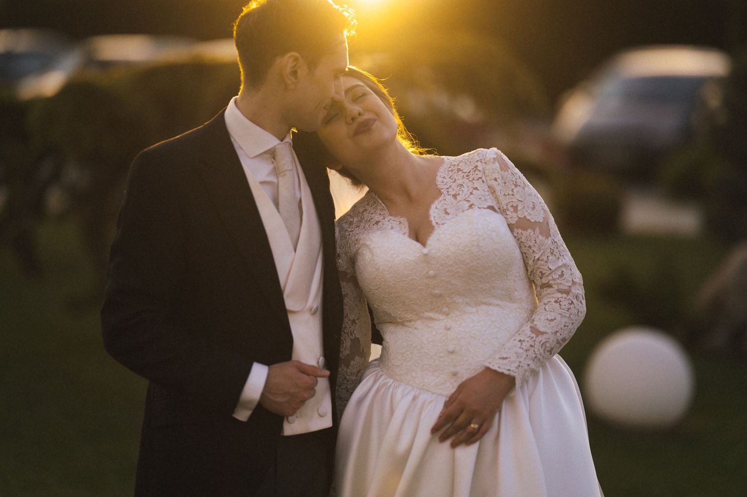 Romantic wedding couple embracing at golden sunset, bride in lace gown and groom in black tuxedo outdoors.