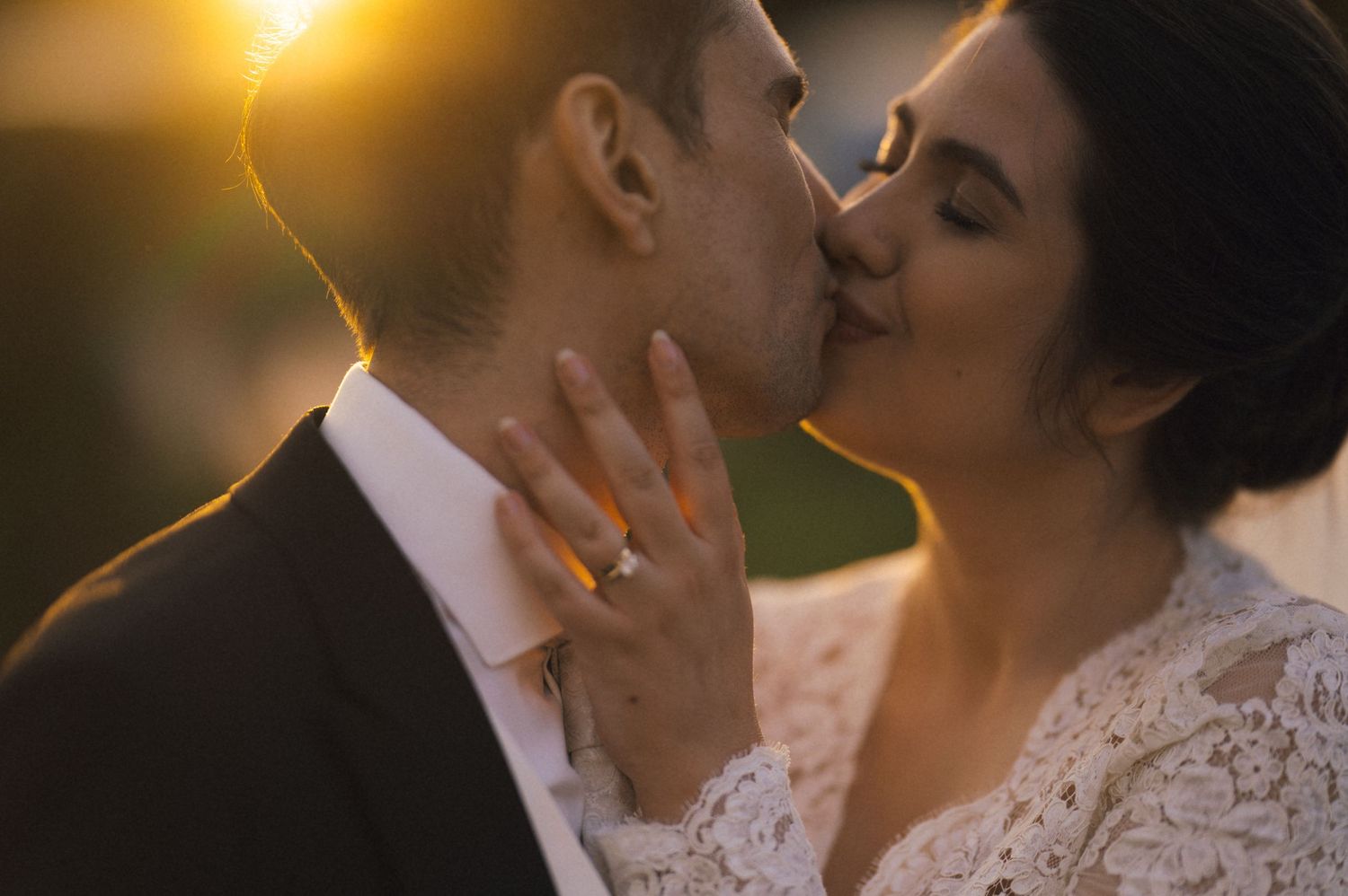 A bride and groom share a romantic kiss at golden hour, her lace dress and diamond ring glowing in the warm sunset light.