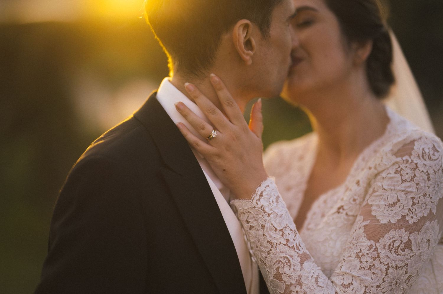 A bride and groom share a romantic kiss at golden hour, with the bride wearing a lace dress and diamond ring.