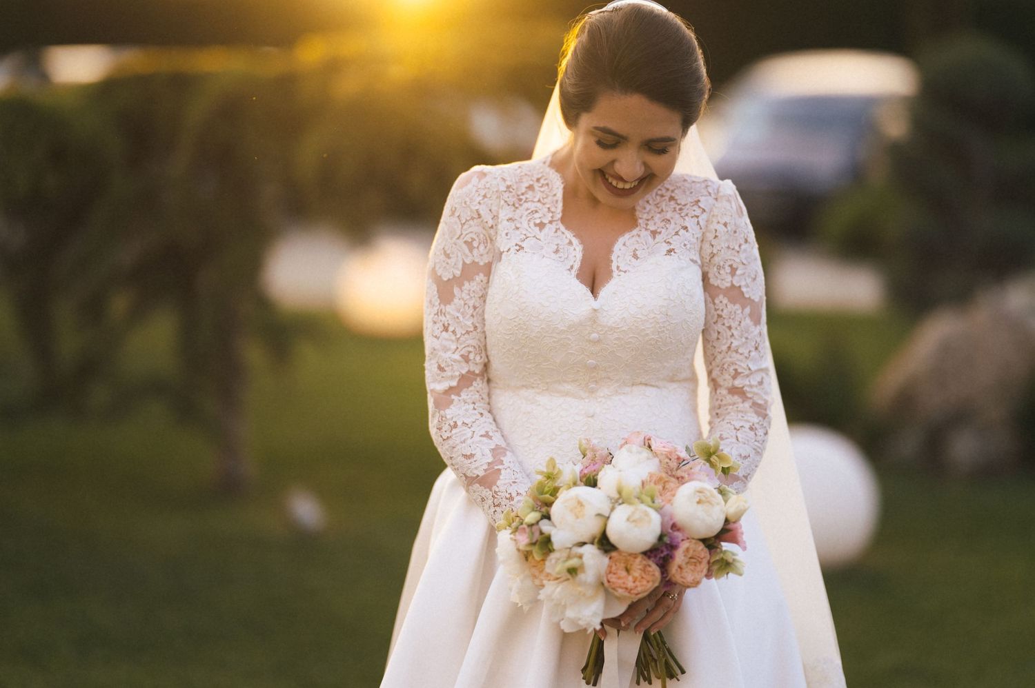 Bride in lace long-sleeve wedding gown smiling while holding a blush pink and white floral bouquet outdoors at sunset.
