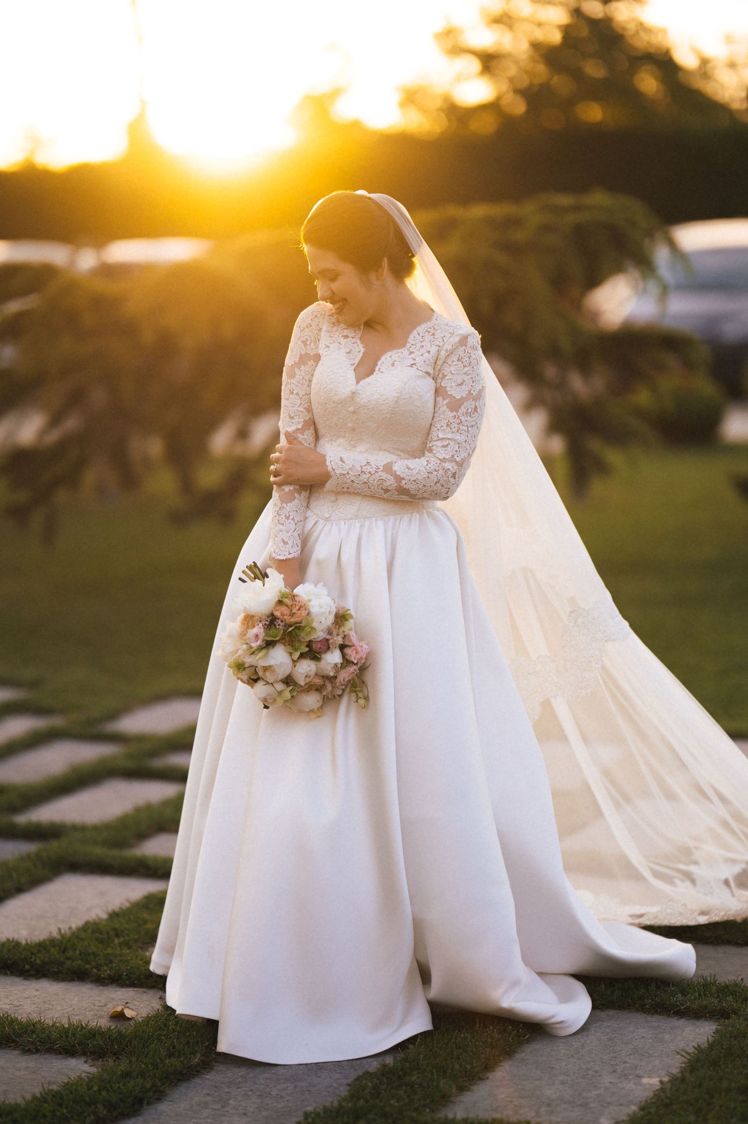 Bride in lace long-sleeve ball gown wedding dress holding floral bouquet at golden hour sunset outdoors.