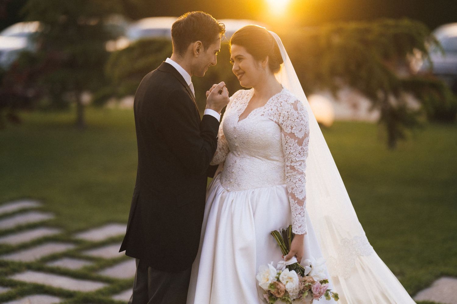 Bride and groom share a romantic sunset moment, holding hands outdoors, bride in lace gown with veil holding a bouquet.