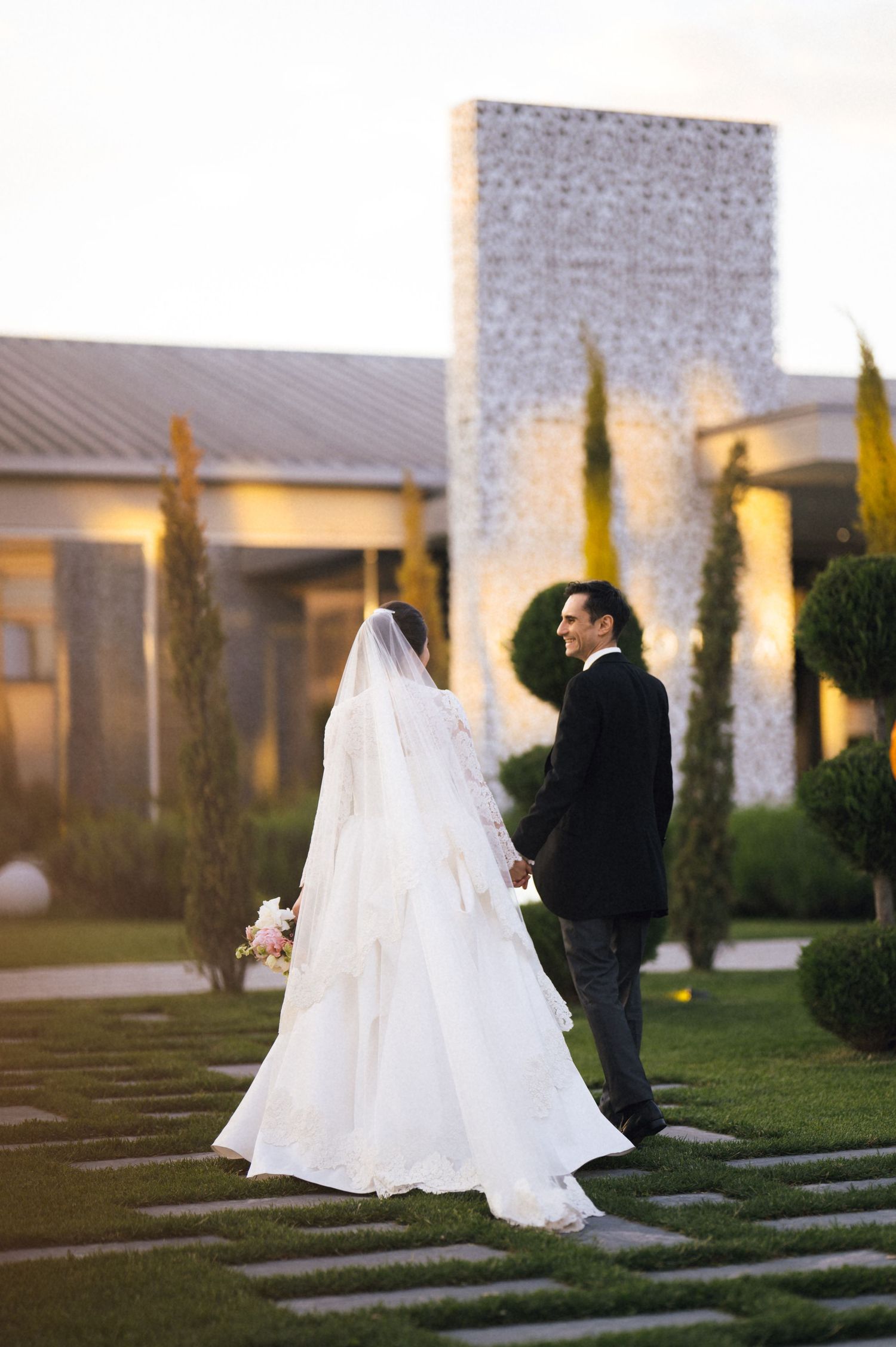 A bride in a white gown and veil walks hand-in-hand with a groom in a black suit through an elegant garden venue.