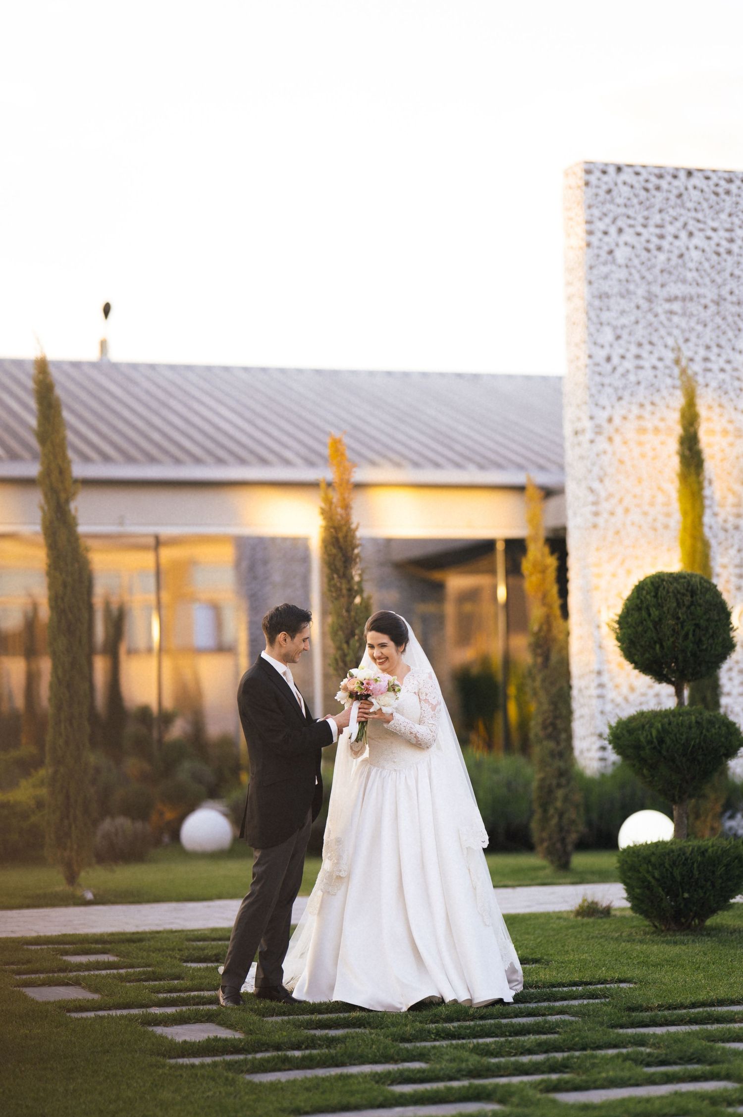 Bride in white gown and groom in black suit stand together in elegant garden venue at sunset.