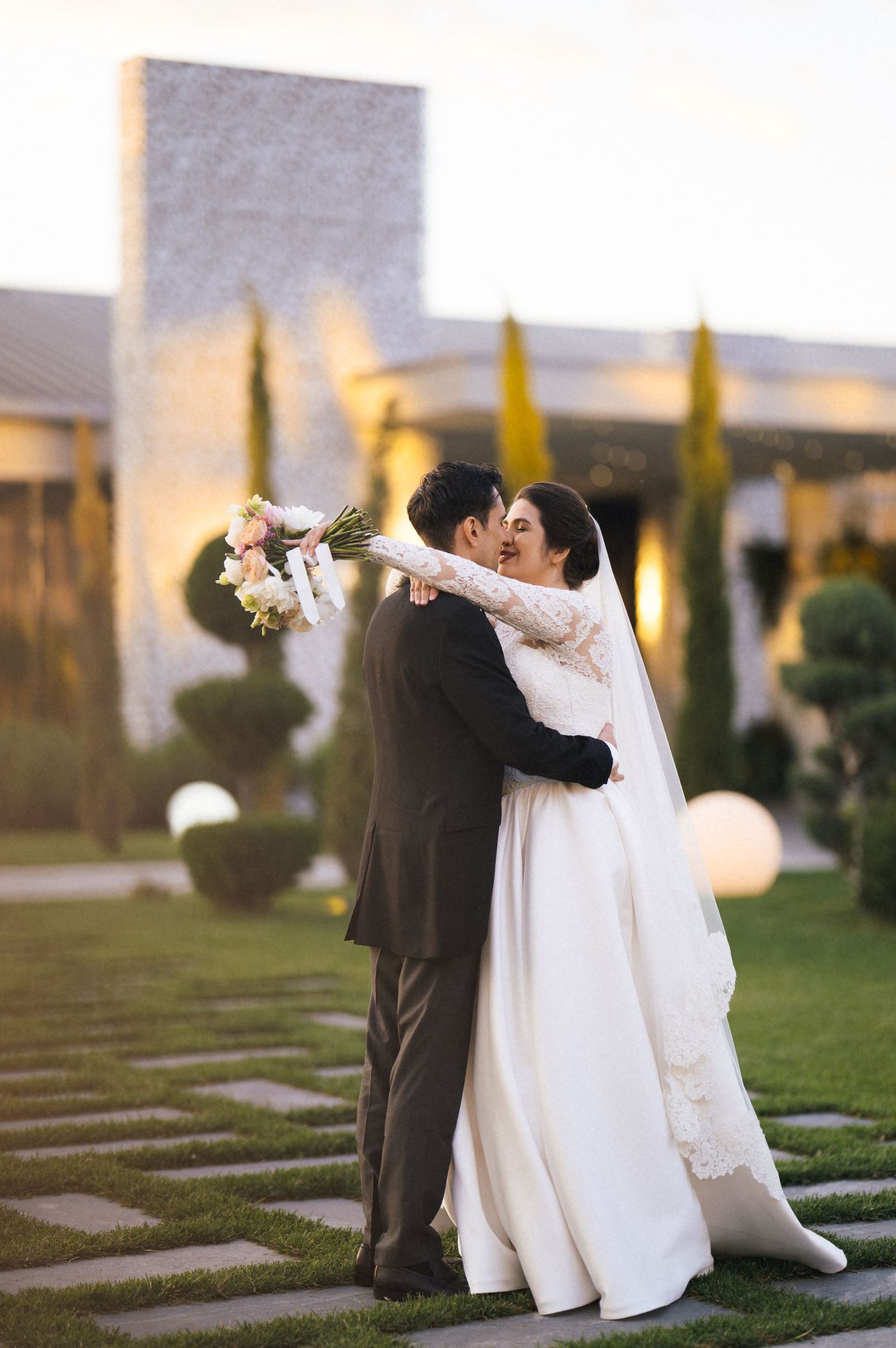 A bride and groom share a romantic kiss outdoors, with the bride holding a floral bouquet in a elegant white gown.