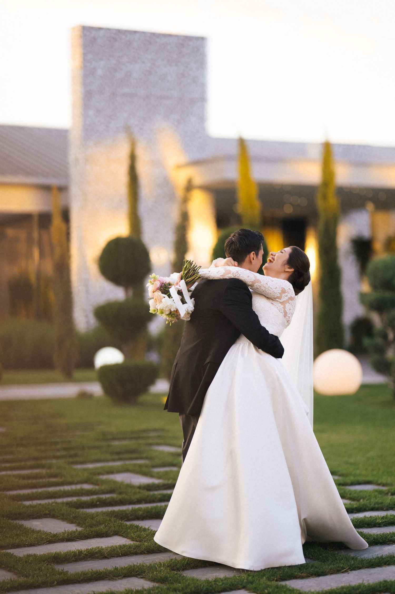 Bride and groom embracing and kissing in a lush garden venue, bride holding white bouquet in elegant lace wedding gown.