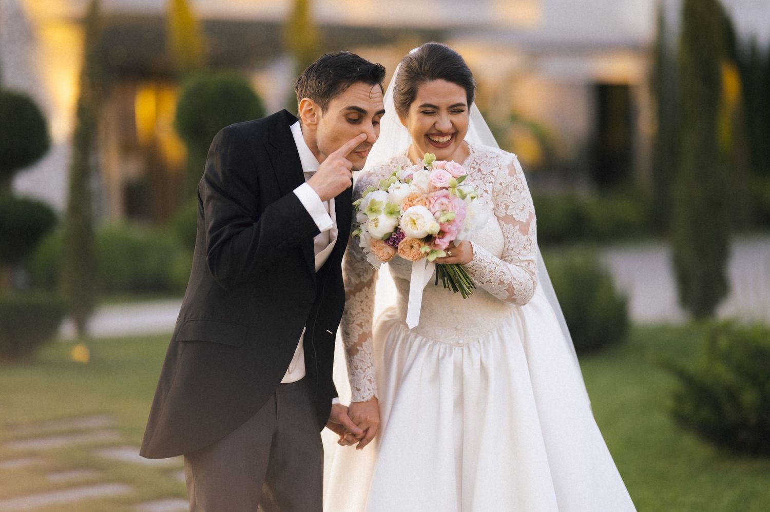 Newlywed couple smiling and holding hands outdoors, bride in lace gown holding bouquet, groom in gray suit.