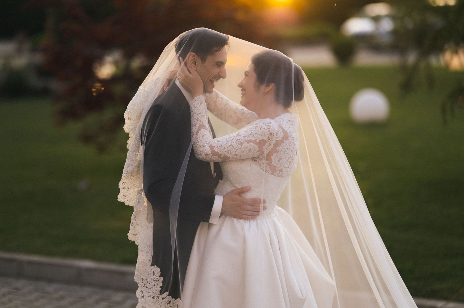 Bride and groom share romantic moment under wedding veil at sunset in outdoor garden ceremony.