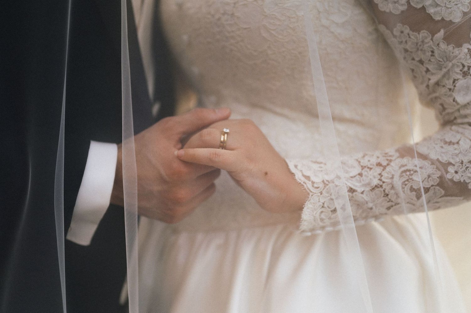 Bride and groom holding hands at wedding ceremony, bride wearing lace dress and gold ring with veil draped over.