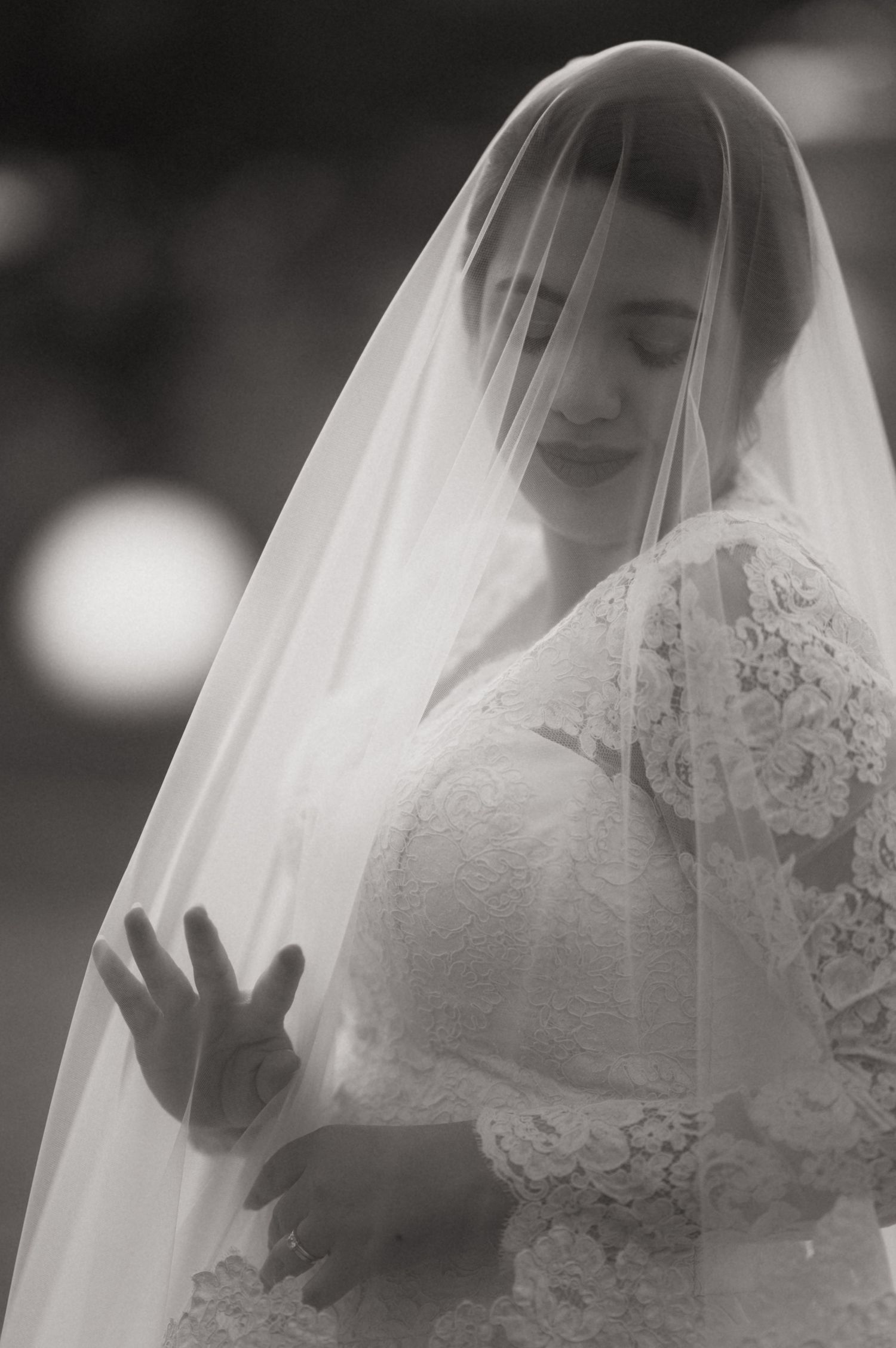 Black and white photo of a bride in lace gown and veil, eyes closed, hands clasped in prayer on her wedding day.
