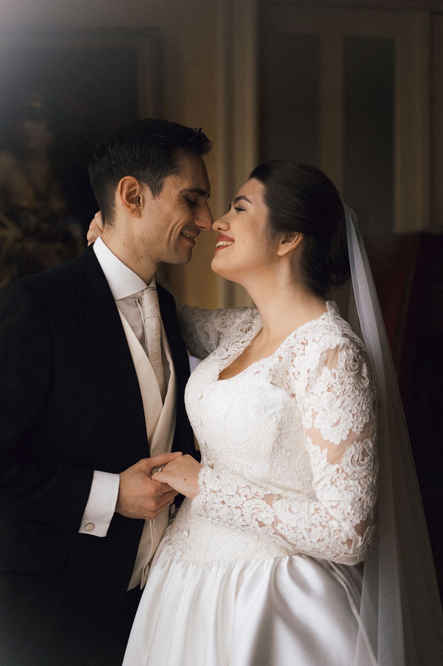 A bride in a lace long-sleeve gown and veil shares a tender moment with her groom in a dark suit on their wedding day.