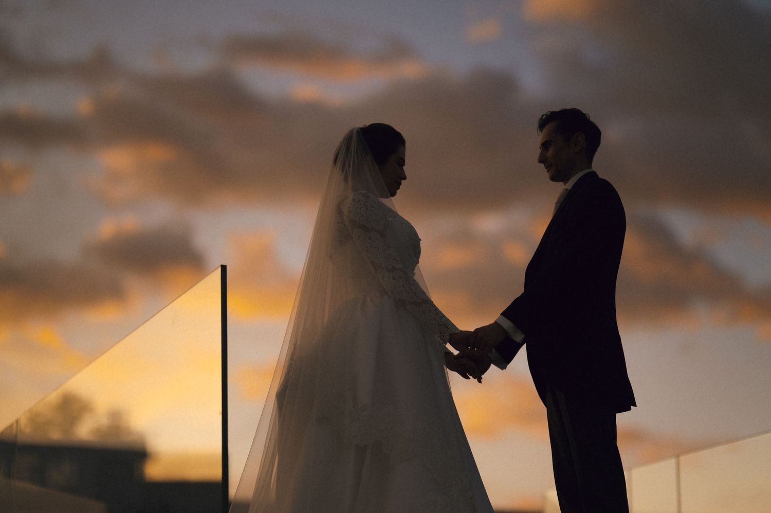 Silhouette of bride and groom holding hands at sunset with dramatic orange and purple cloudy sky backdrop.