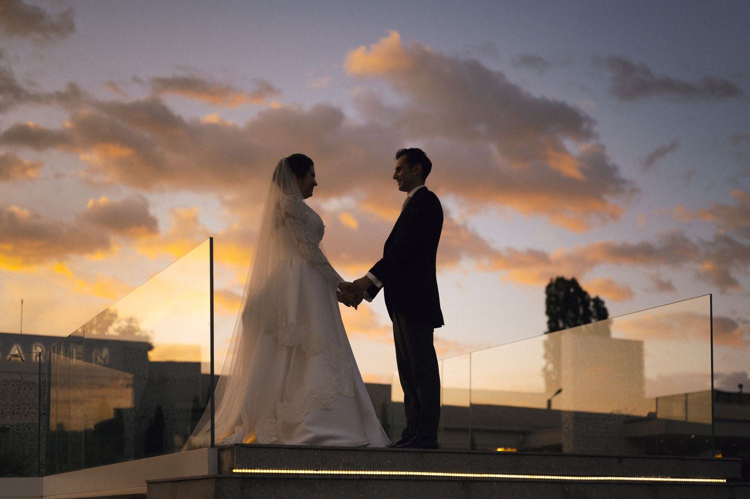 Bride and groom holding hands at sunset on a rooftop, silhouetted against a dramatic orange and purple sky.