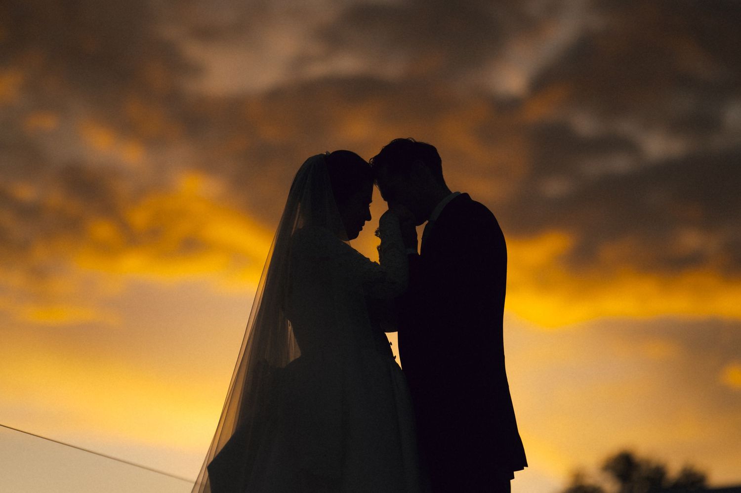 Silhouette of bride and groom facing each other against a dramatic golden sunset sky on their wedding day.