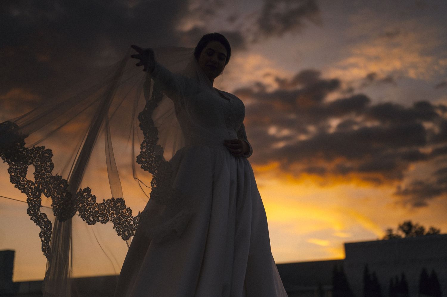 Bride in white gown and veil poses dramatically against a stunning golden sunset sky with dramatic clouds.