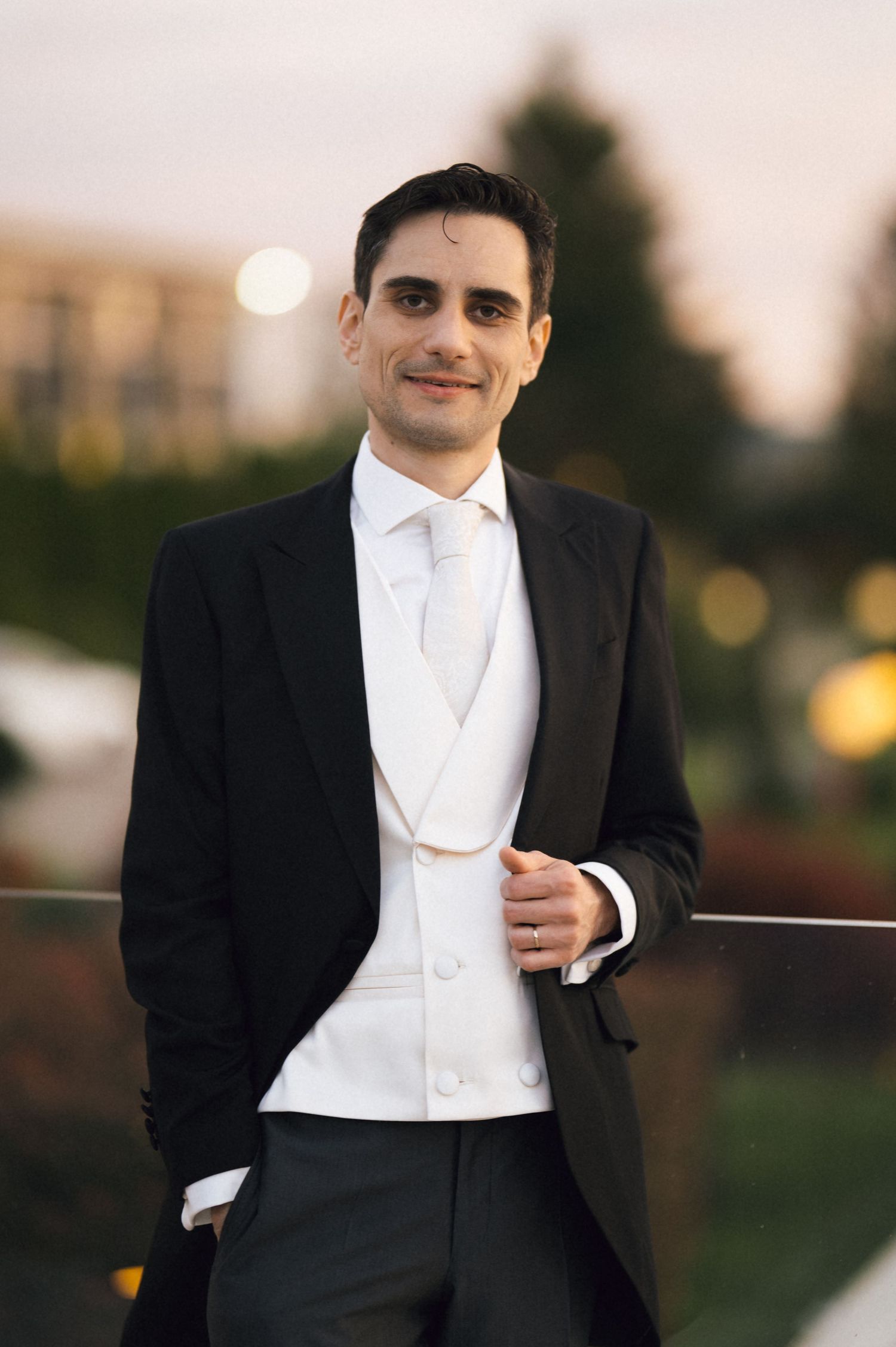 Elegant groom in black tuxedo with white vest and tie smiling outdoors at dusk.