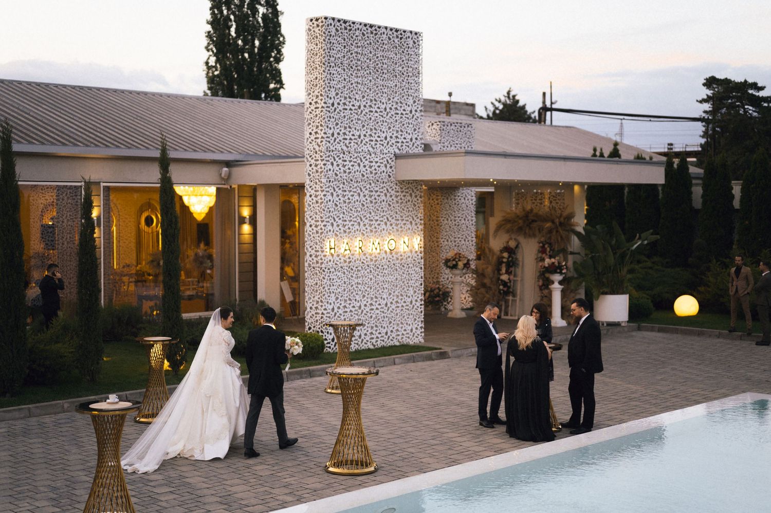 Elegant outdoor wedding ceremony by a pool at dusk, bride in white gown with groom and guests near decorative wall.